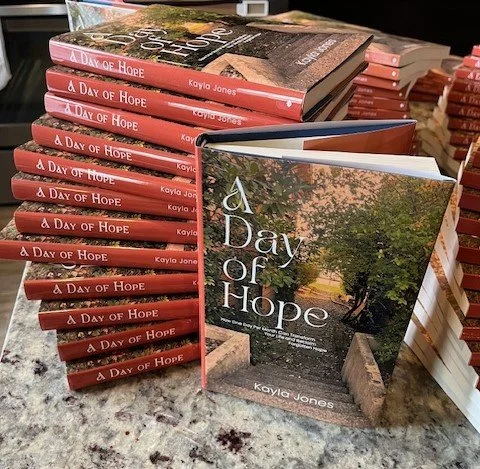 A stack of hardcover books titled 'A Day of Hope' by Kayla Jones, displayed on a table. One copy is open showing the cover with a tree and a sunrise scene, suggesting it is a published book.