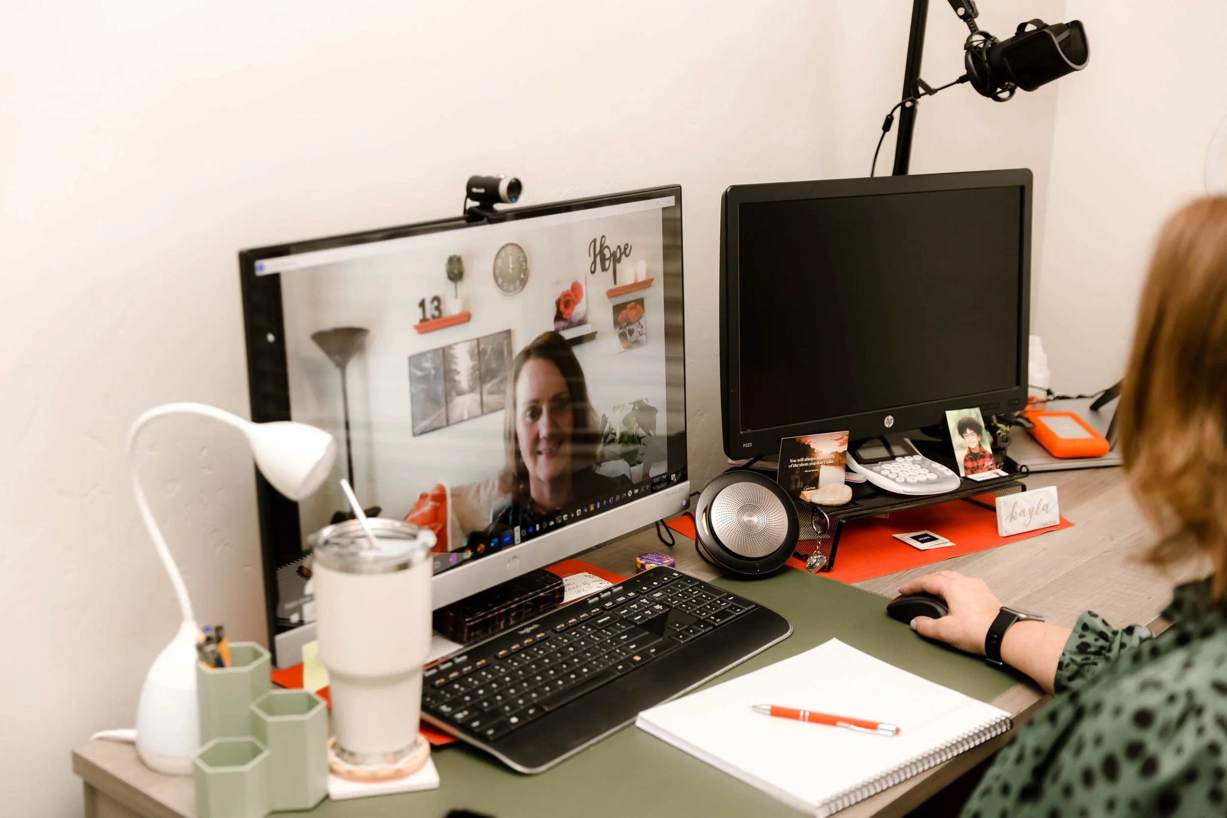 Desk setup with two computer monitors, one displaying a woman on a video call, on a desk with notebooks, pens, a water bottle, and various office supplies.