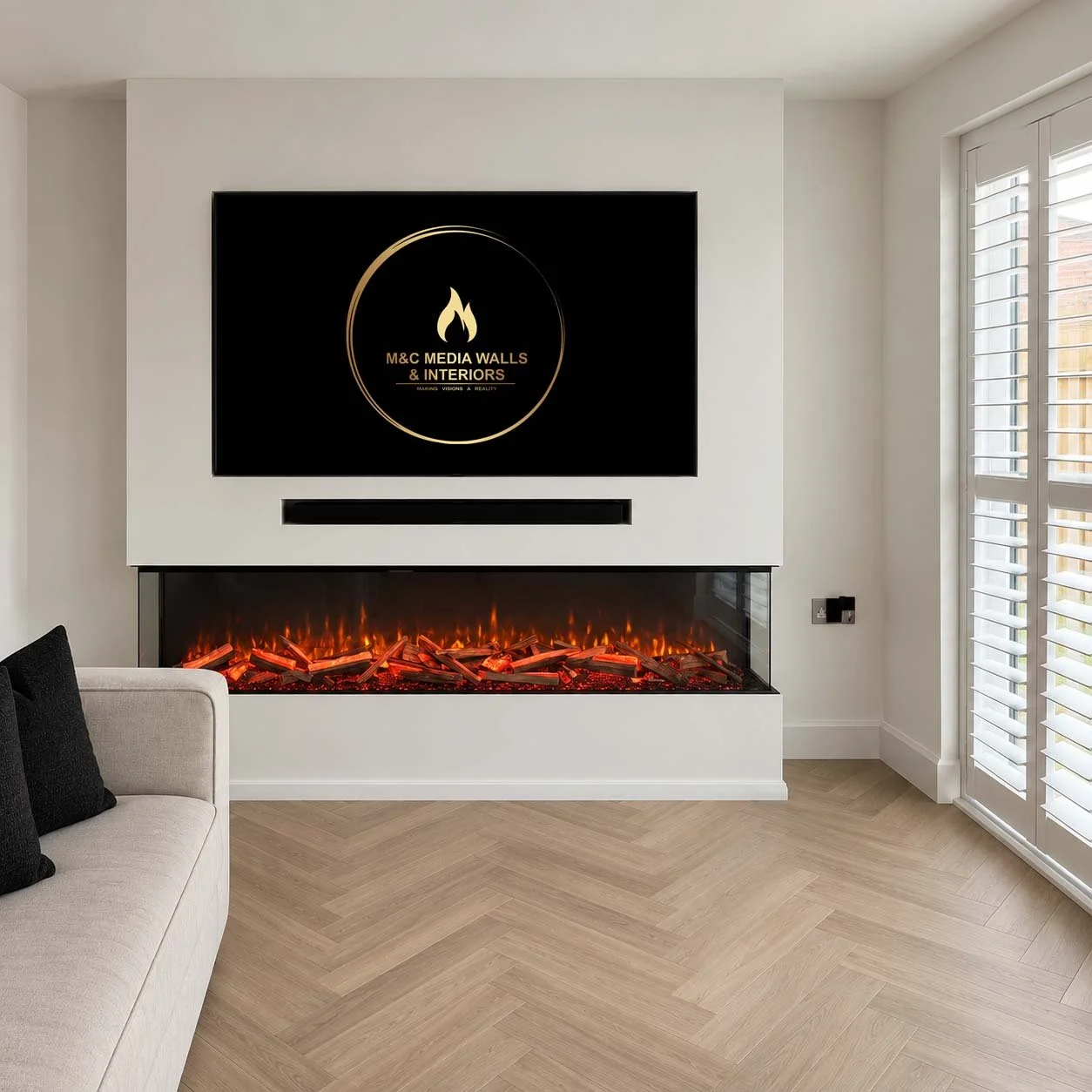 Modern living room with a white wall-mounted electric fireplace and a large flat-screen TV above it. The TV screen displays a logo for M&C Media Walls & Interiors. To the right, there are glass doors with white shutters, and on the left, part of a light-colored sofa with black pillows. The room has light wooden flooring.