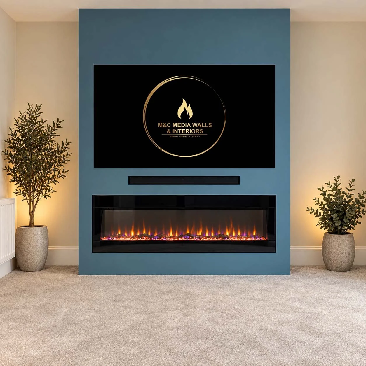 Living room with beige carpet, two potted plants flanking a modern electric fireplace, and a large wall-mounted television displaying M&C Media Walls & Interiors logo with a flame icon.