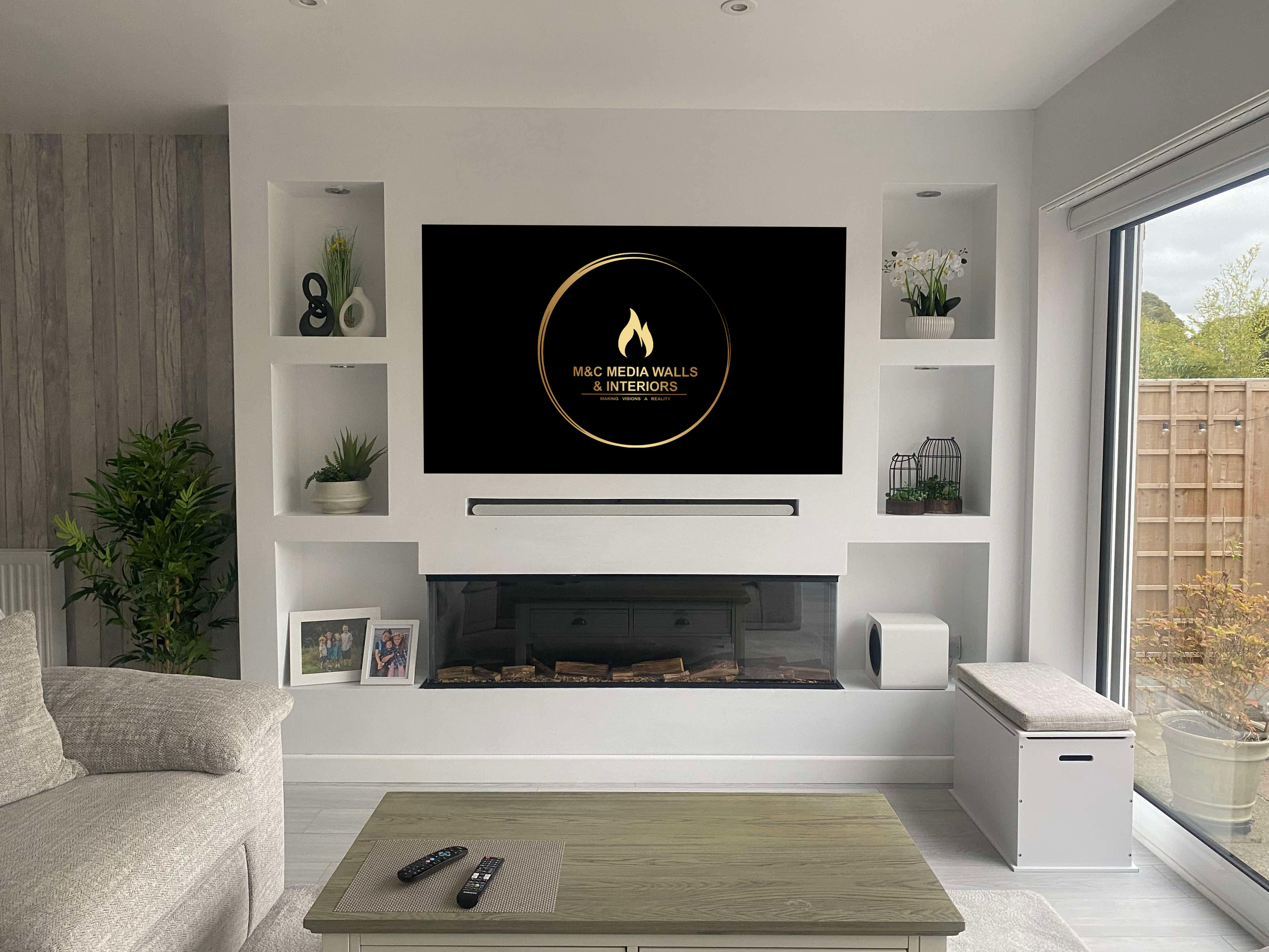 Living room with a white wall, built-in shelves, and a fireplace. A large flat-screen TV displays the logo for M&C Media Walls & Interiors. Decor includes plants, framed photos, and birdcages. A wooden coffee table with remotes is in the foreground; a beige sofa is on the left, and a sliding glass door revealing an outdoor fence and trees is on the right.