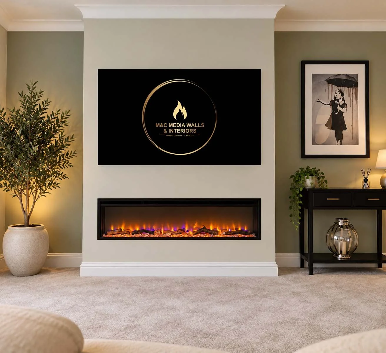Living room with a fireplace, a large flat-screen TV displaying a logo for M&C Media Walls & Interiors, a potted plant to the left, and a black console table with a plant, lamp, and decorative jar on the right; framed art on the wall.