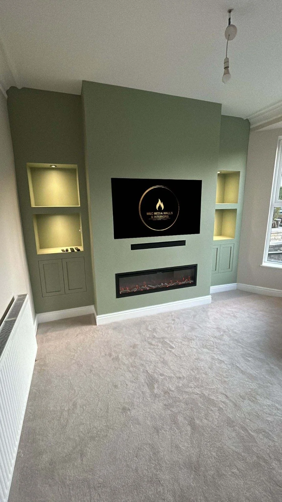 Modern living room with a glass coffee table, an electric fireplace, and a wall-mounted screen showing the logos of "M&C Media Walls" and "Bespoke Fireplaces."