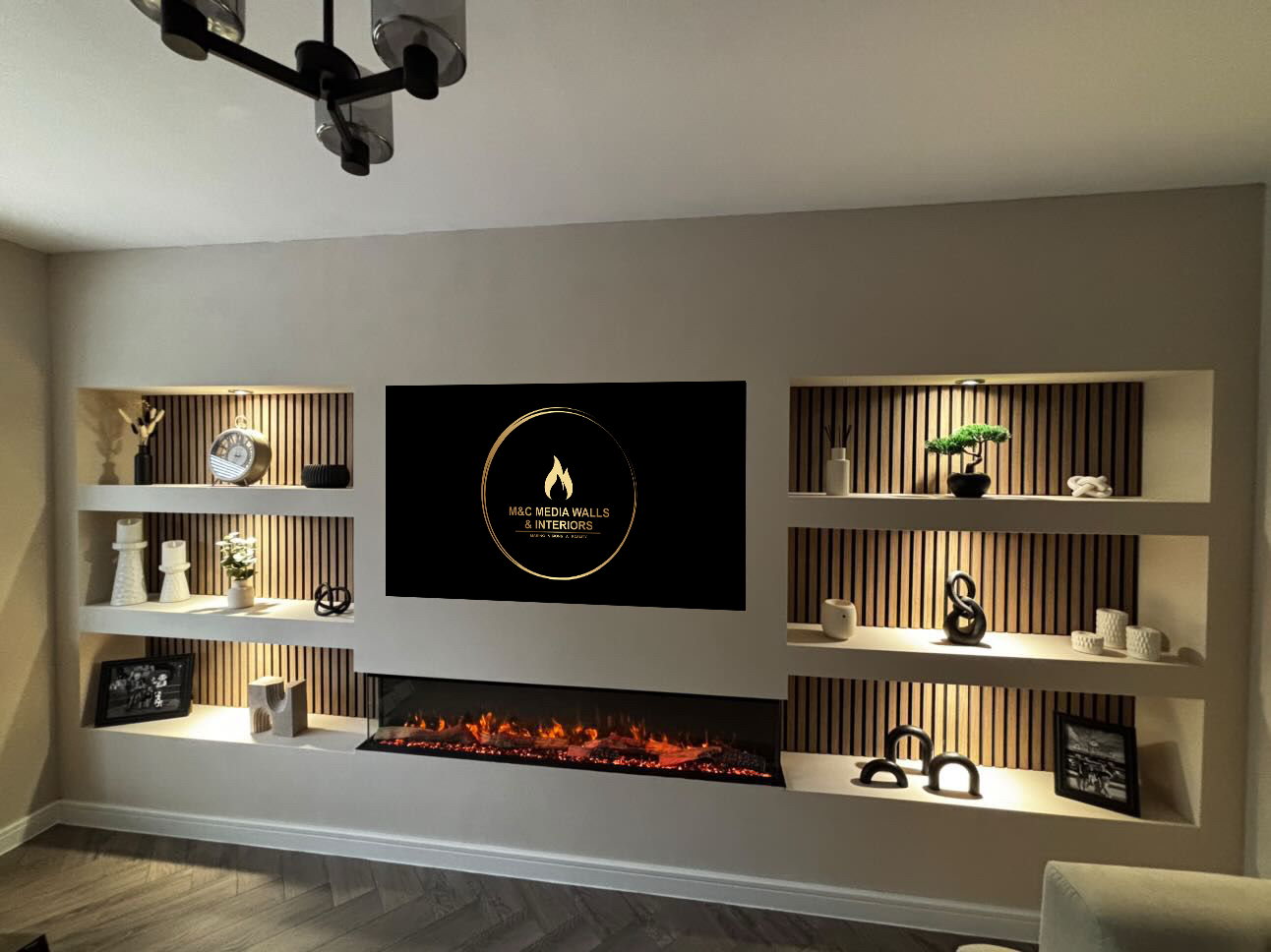 Living room wall with built-in shelves, decorative items, a digital fireplace, and a television displaying the logo for M&C Media Walls & Interiors.