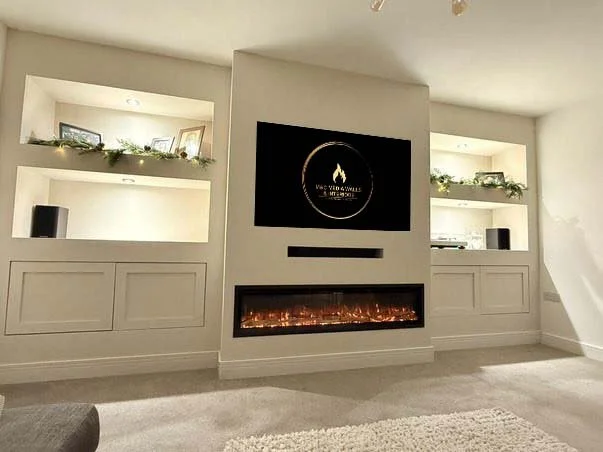 Modern living room with custom built-in shelving, a large TV screen displaying "Bespoke Fireplaces," and a contemporary electric fireplace beneath it. The carpeted floor and recessed lighting provide a cozy ambiance.