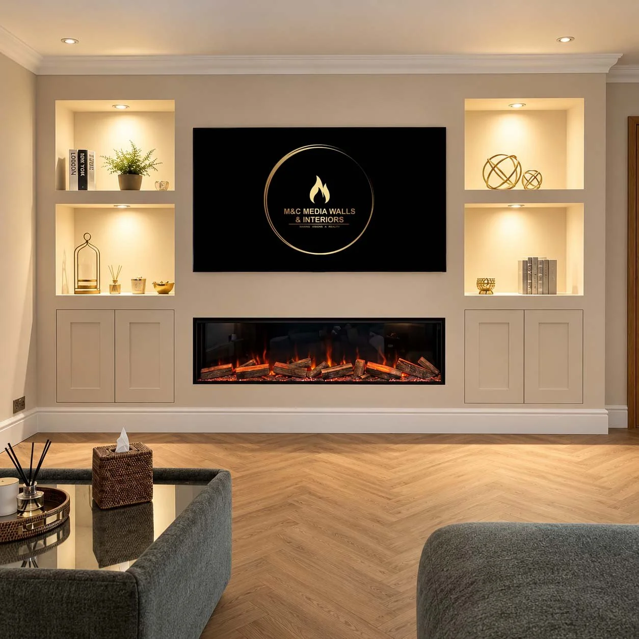 Living room with a wall-mounted television displaying 'M&C Media Walls & Interiors' logo, a modern electric fireplace underneath, built-in illuminated shelves with decorative items, a dark sofa, and a wooden floor.