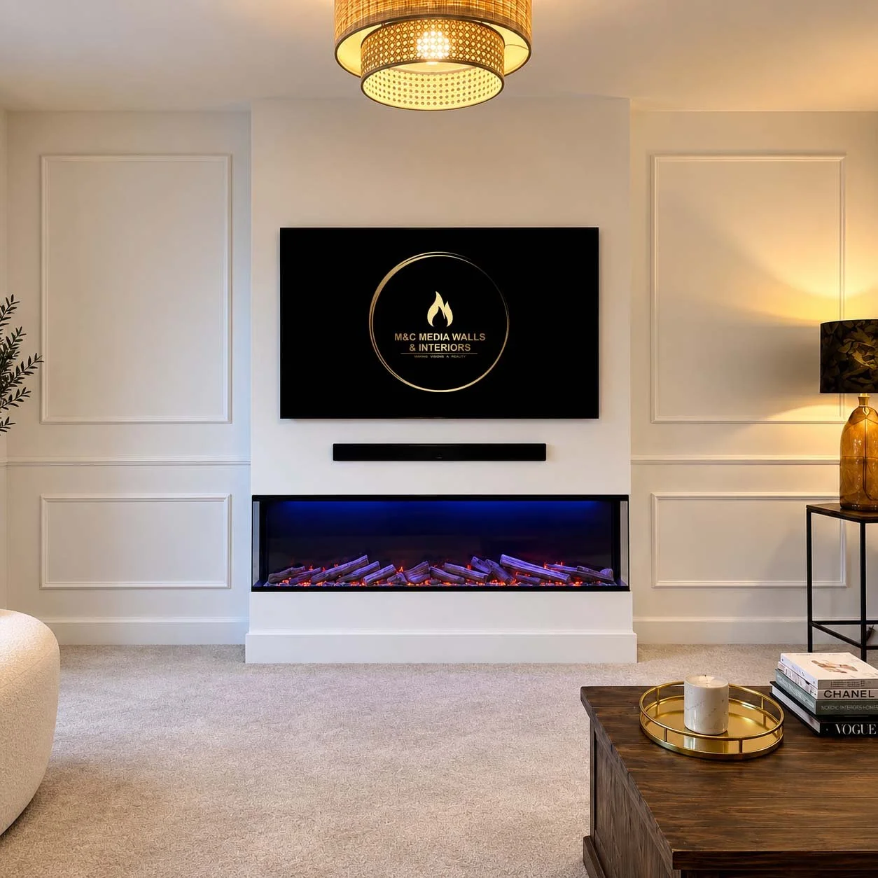 Living room with a modern electric fireplace below a wall-mounted TV displaying a logo for 'M&C Media Walls & Interiors.' The room is decorated with white paneled walls, a black table lamp, a wooden coffee table with books, and a large gold vase, all illuminated with warm lighting.