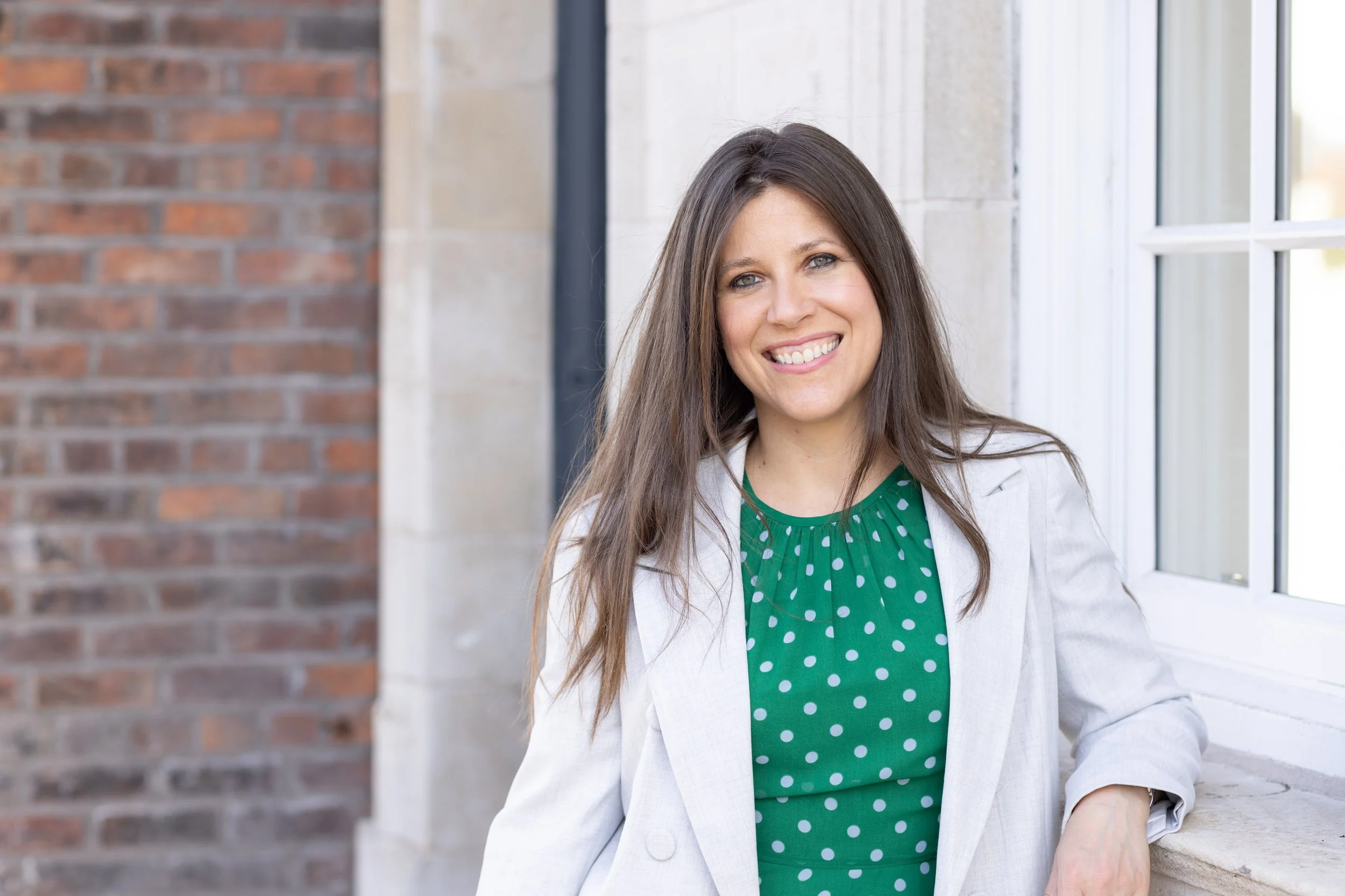 A smiling woman with long brown hair, wearing a white blazer over a green polka dot shirt, standing near a window outdoors.