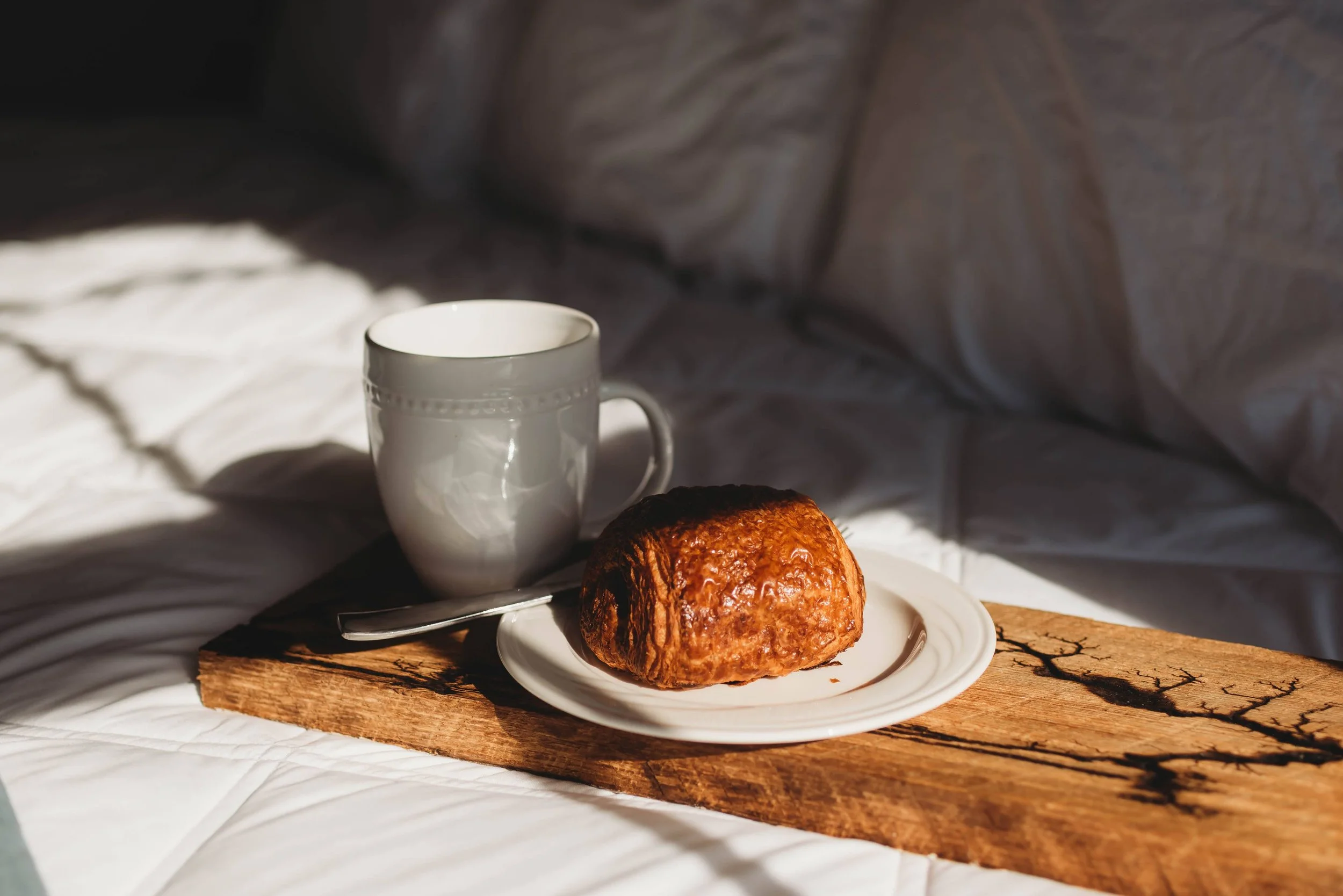 A flaky pastry and coffee sit on a wooden tray atop a bed in soft morning light, capturing a relaxed brunch the day after a wedding at a venue with lodging. Crisp white bedding and warm sunlight create a cozy post celebration atmosphere.