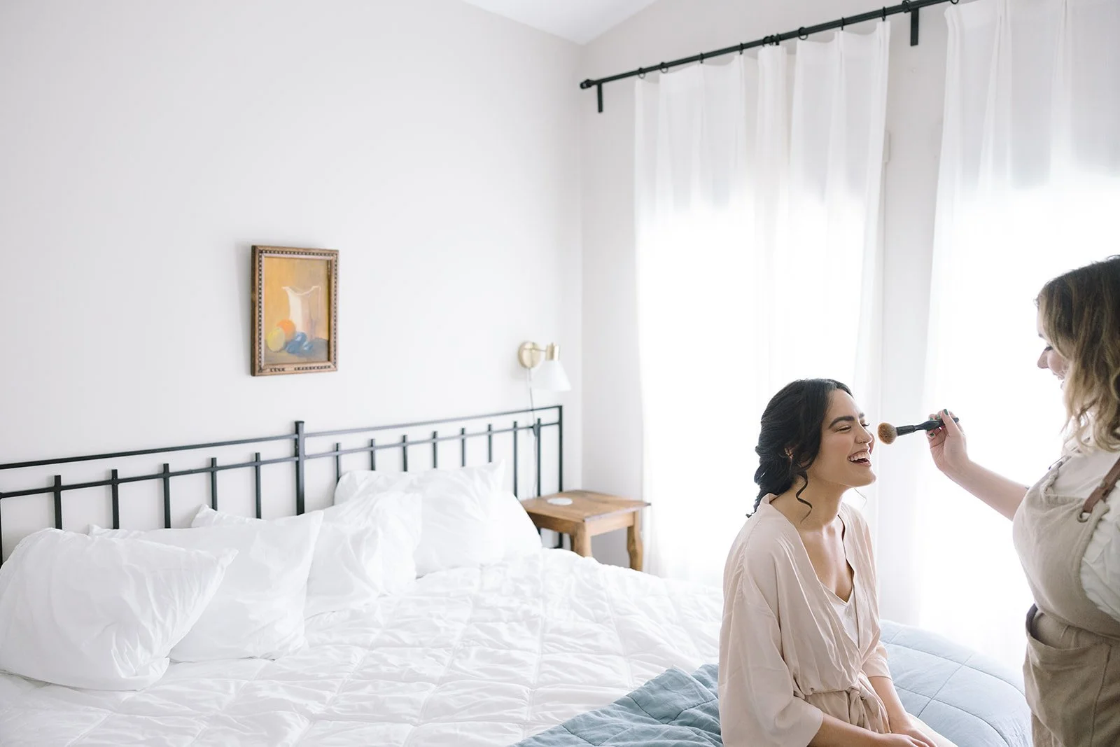 Bride sits on the edge of a bed in a bright suite at a wedding venue with lodging while a makeup artist applies blush to her face. Soft natural light through sheer curtains highlights a calm getting ready moment before the ceremony.