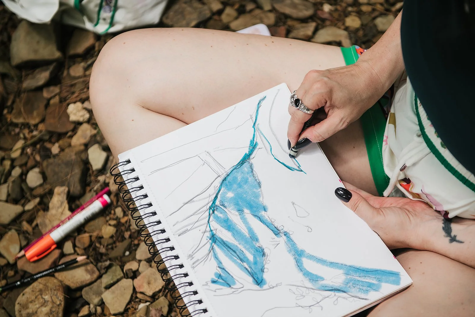 Close up of a person sketching a blue landscape in a notebook while sitting outdoors, highlighting a creative retreat activity in nature.