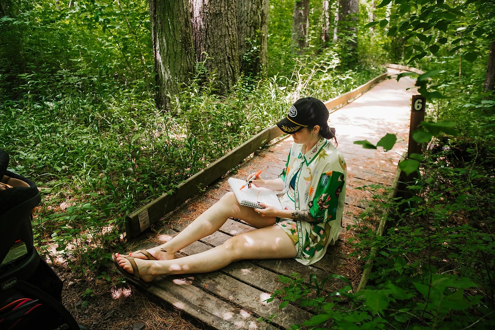A woman sits on a forest path sketching in a notebook surrounded by lush greenery. Text above reads