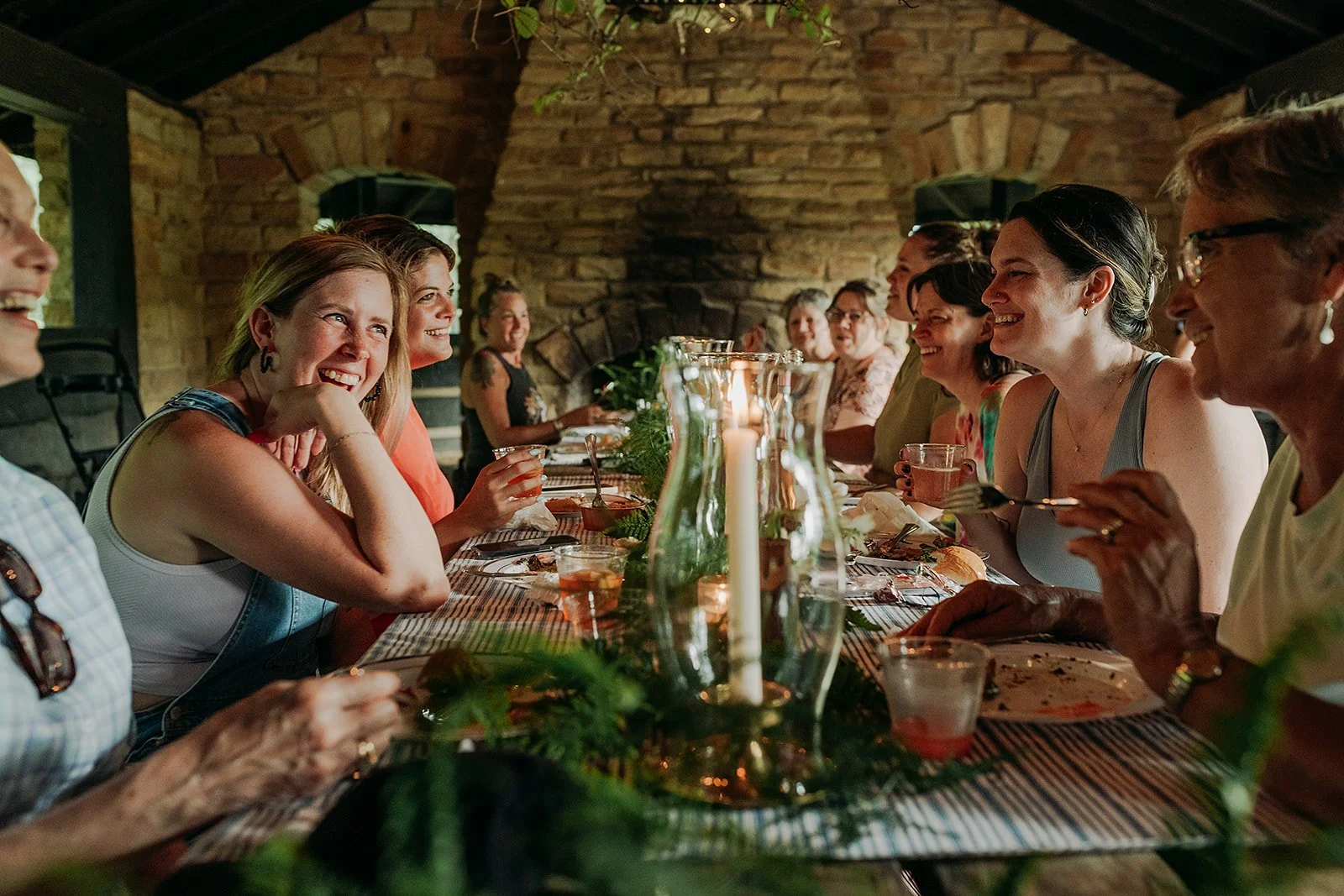 People laugh and share a meal together around a long table in a cozy candlelit stone space, creating a warm and connected retreat experience.