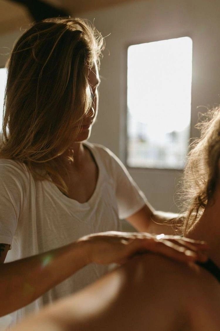 A woman with long hair and a white shirt giving a massage in a room with a window letting in sunlight.