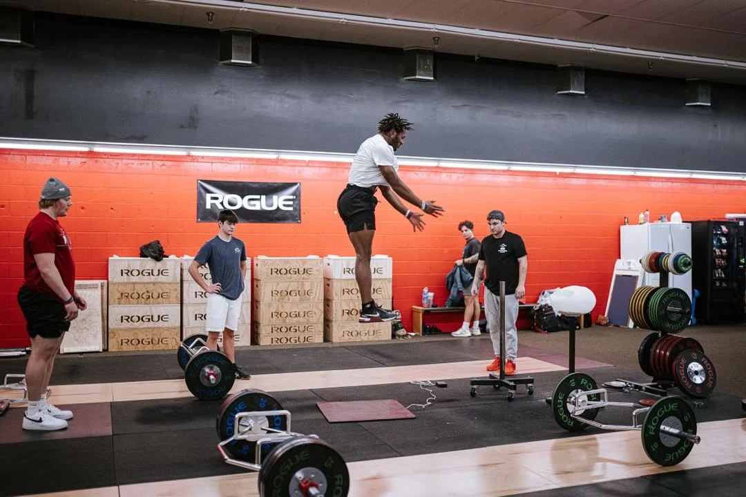 A person jumps onto a elevated platform in a gym with weightlifting equipment and four other people watching.