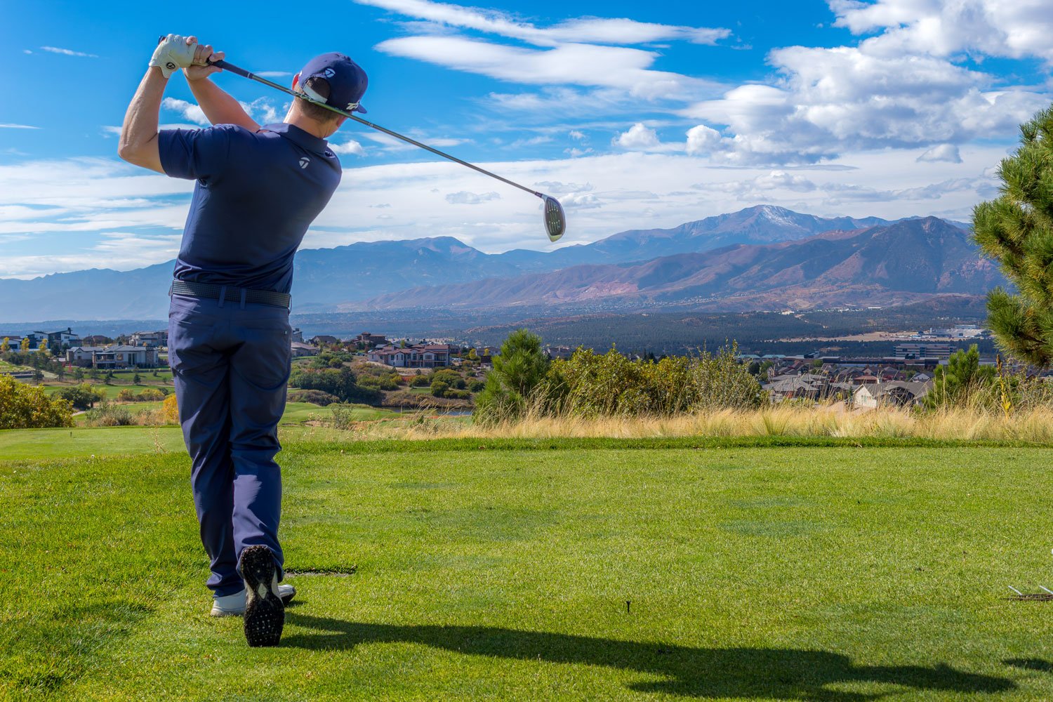 A man in a navy golf shirt, pants, and cap swings a golf club on a golf course with mountains in the background.