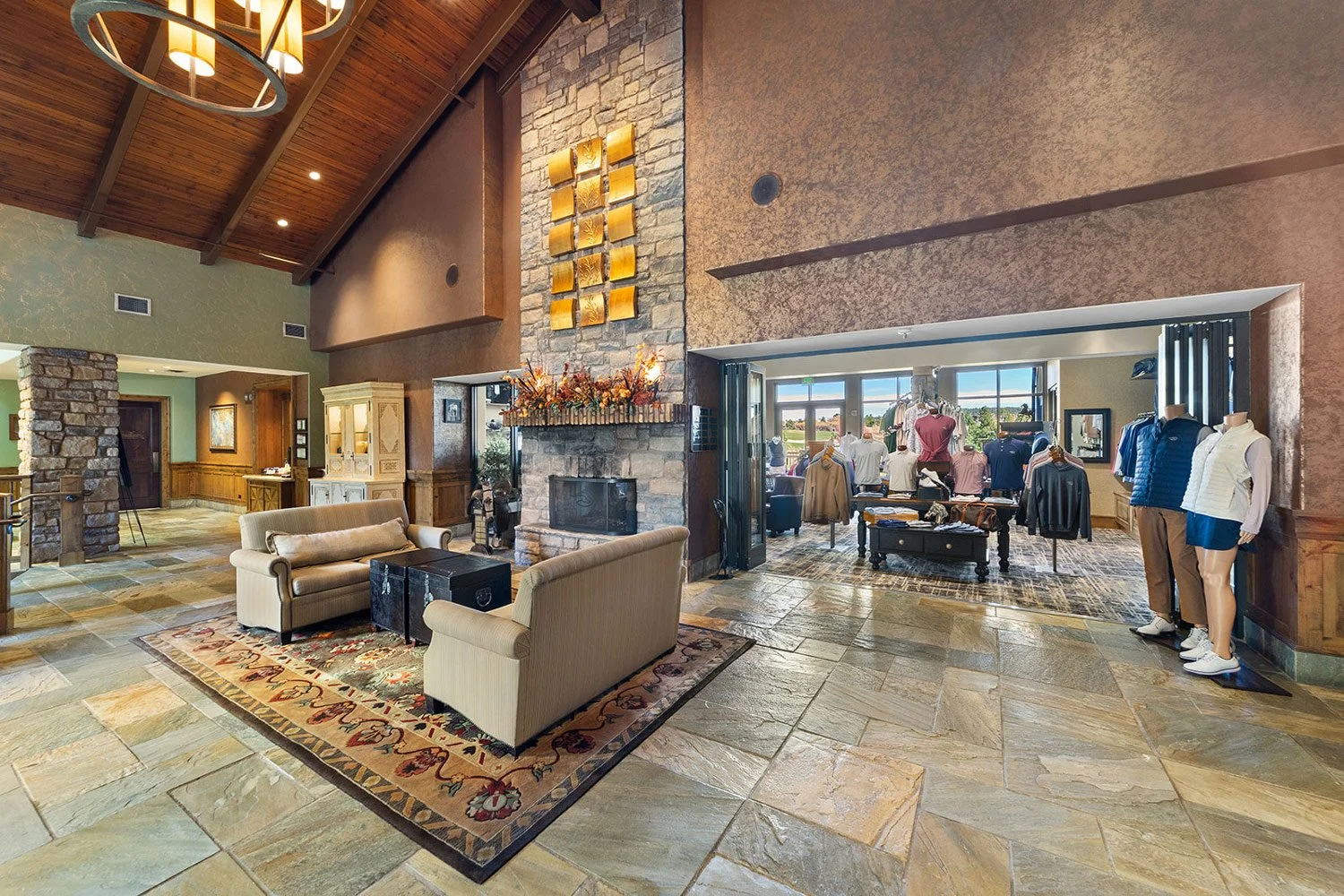 Hotel lobby with seating area, fireplace, stone walls, and a clothing store with mannequins displaying golf apparel near large windows.