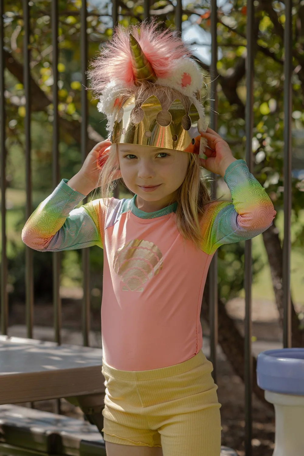 A young girl in colorful, rainbow-patterned long sleeves and a pink shirt stands outdoors, adjusting a shiny gold unicorn horn headpiece adorned with pink and white fluffy material and a pink feather. She looks at the camera with a playful expression