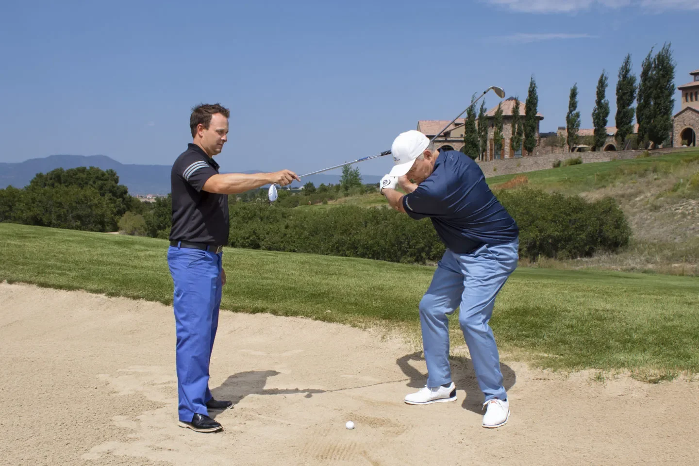 A golfer taking a shot from a sand bunker on a golf course while another man stands nearby, watching.