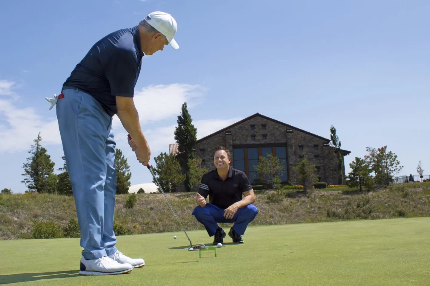 Two men on a golf course, one is preparing to putt, and the other is squatting and giving instructions. There is a house and trees in the background under a blue sky.