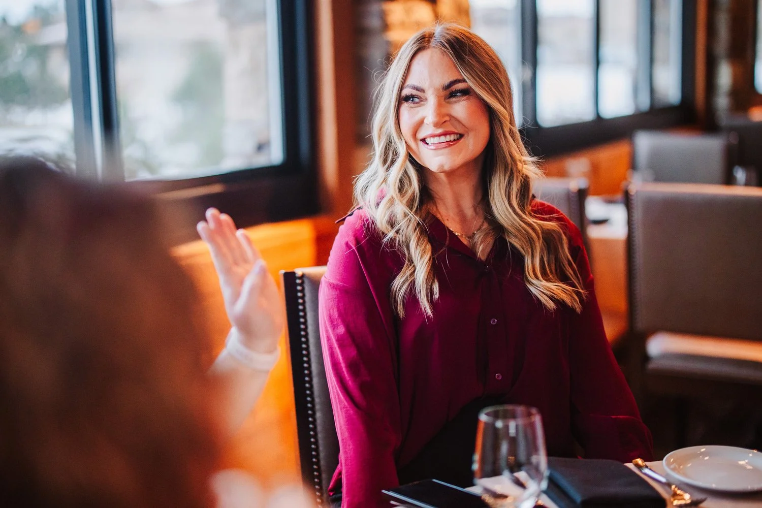 A woman with long, wavy blonde hair and a red blouse smiling and engaging in conversation at a restaurant table with a glass of wine, plate, and utensils visible.