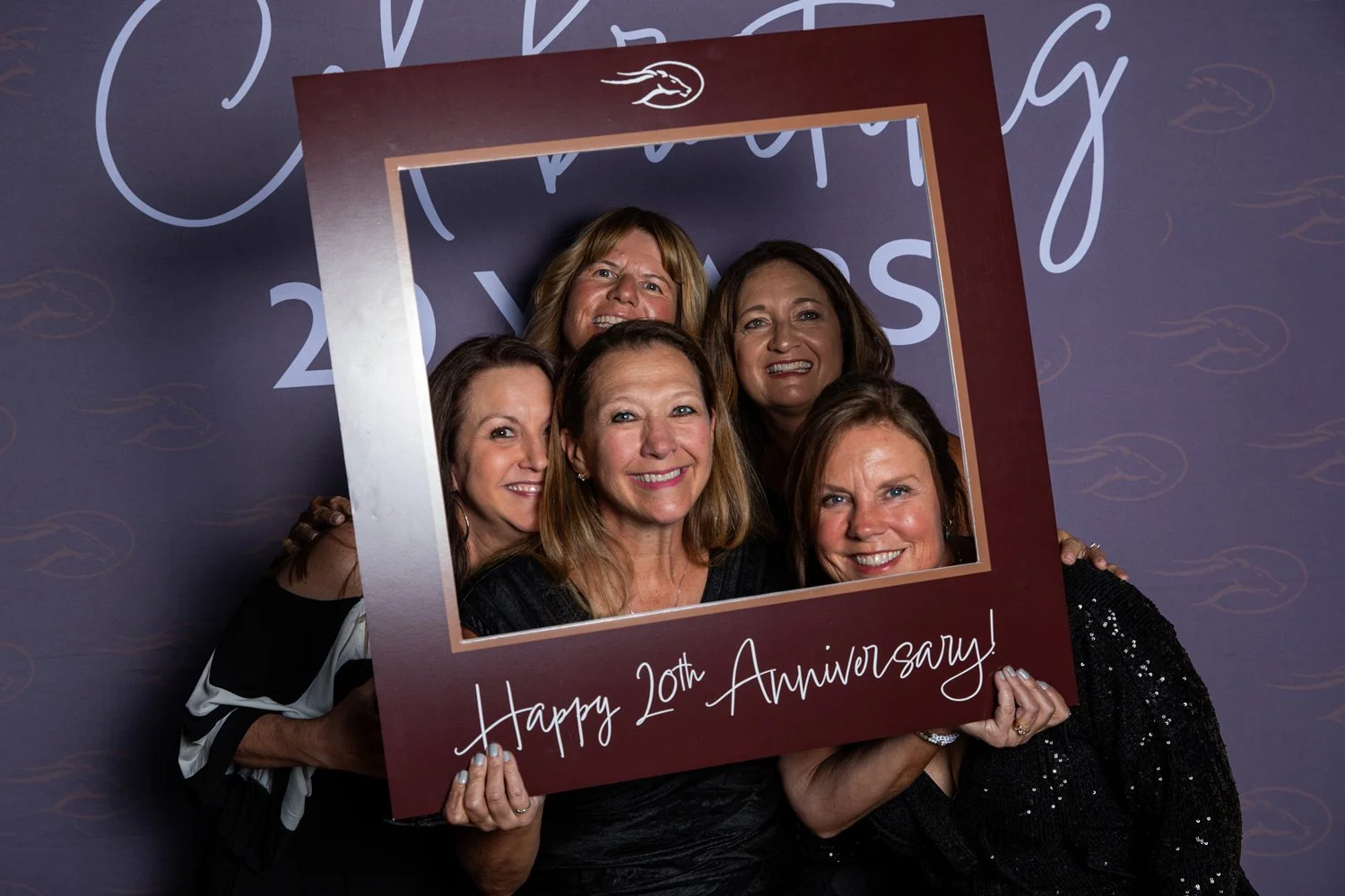 Group of six women smiling and holding a photo frame that says 'Happy 20th Anniversary!' at a celebration event.