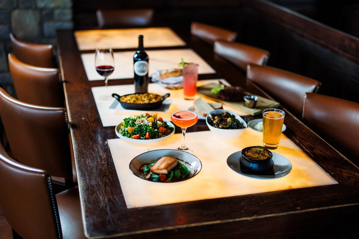 A restaurant table set with various dishes, drinks, and a wine bottle, all arranged on a glowing white table surface, surrounded by brown leather chairs.