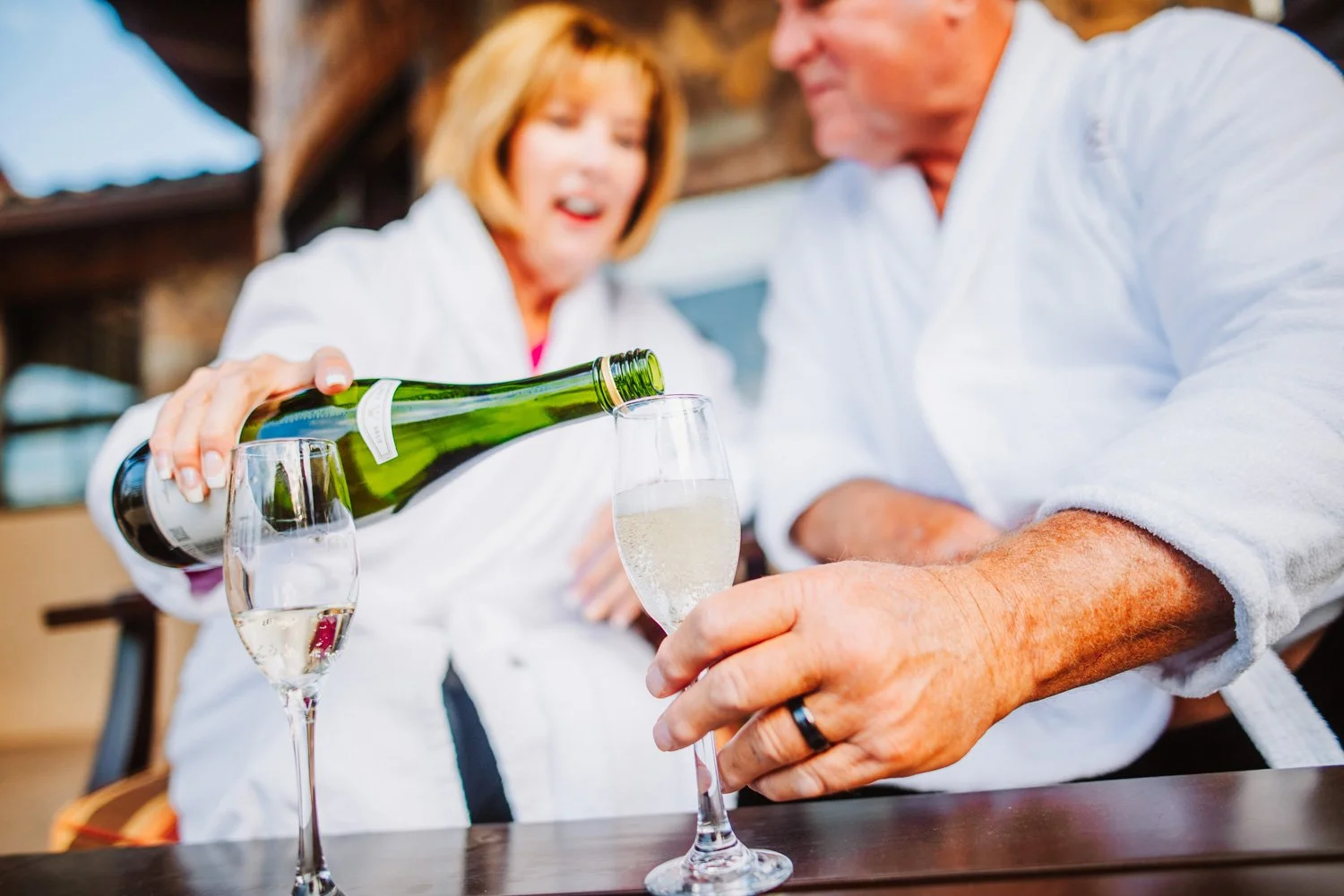 A man and woman clink glasses of champagne outdoors, with the woman pouring bubbly into her glass.