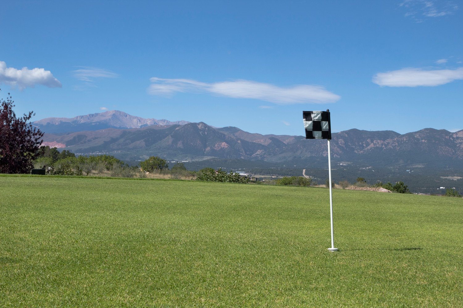 A golf course with a green fairway and a black & white checkered flag against a backdrop of mountains and a partly cloudy blue sky.