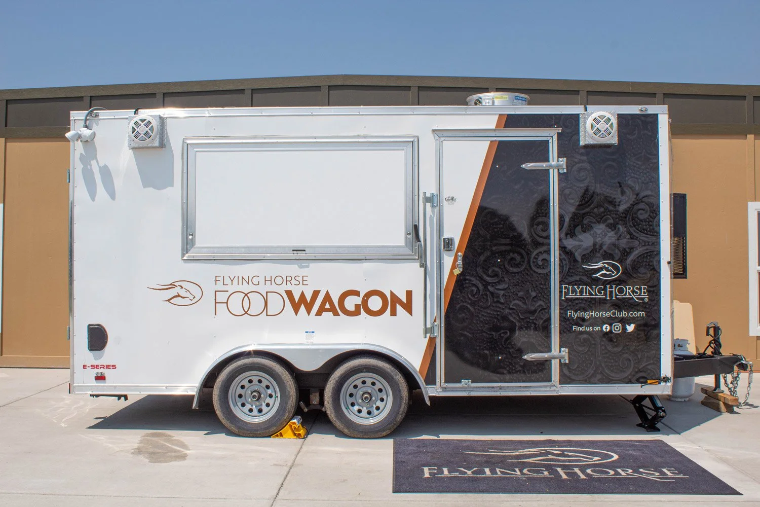 A food truck named 'Flying Horse Food Wagon' parked on a concrete surface with a brown building in the background. The truck is white with a black and brown design on the right side and logos, including social media icons, on the lower right side.