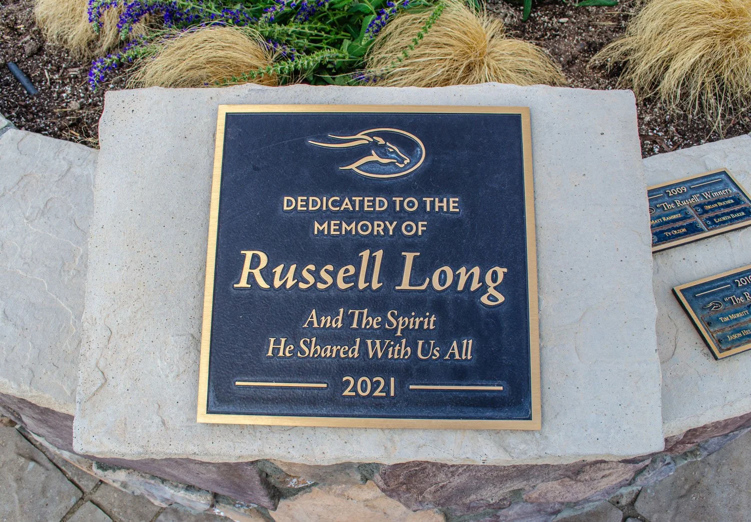 A memorial plaque by the pavilion dedicated to Russell Long, surrounded by landscaping rocks and plants.