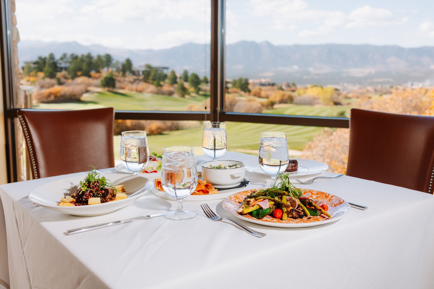 A table set for three with plates of pasta and vegetables, four glasses of water, and a bowl of soup, overlooking a scenic landscape of mountains and trees through large windows.