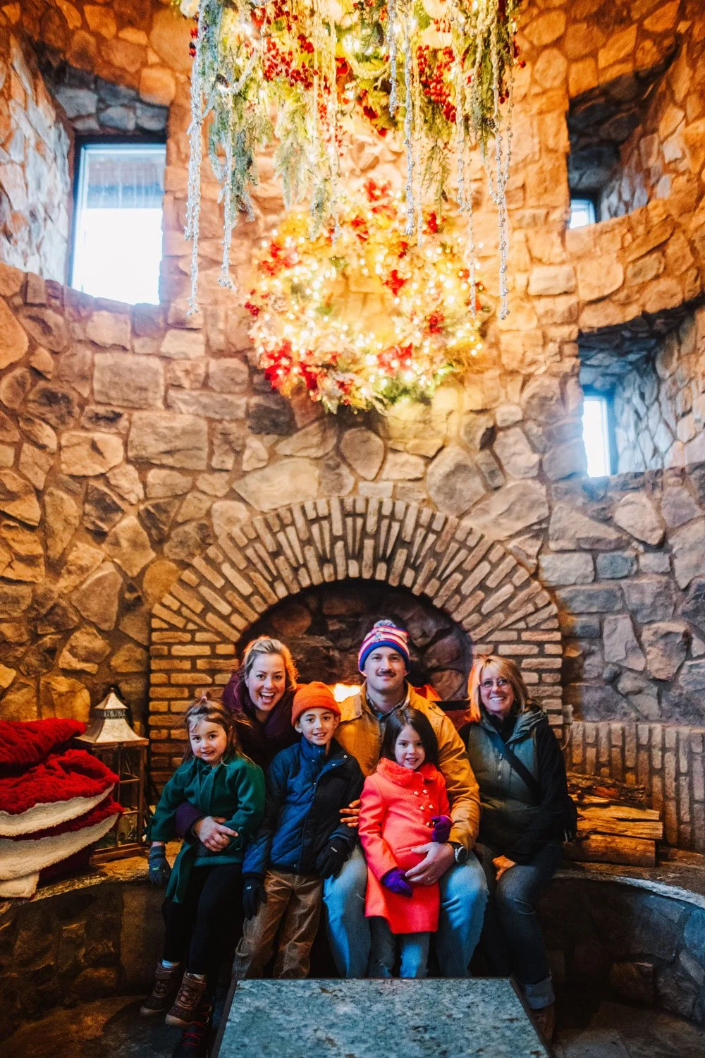 Family of five sitting in front of a large stone fireplace decorated with Christmas ornaments, with a Christmas wreath hanging above and Christmas banners on the side, inside a cozy stone room.