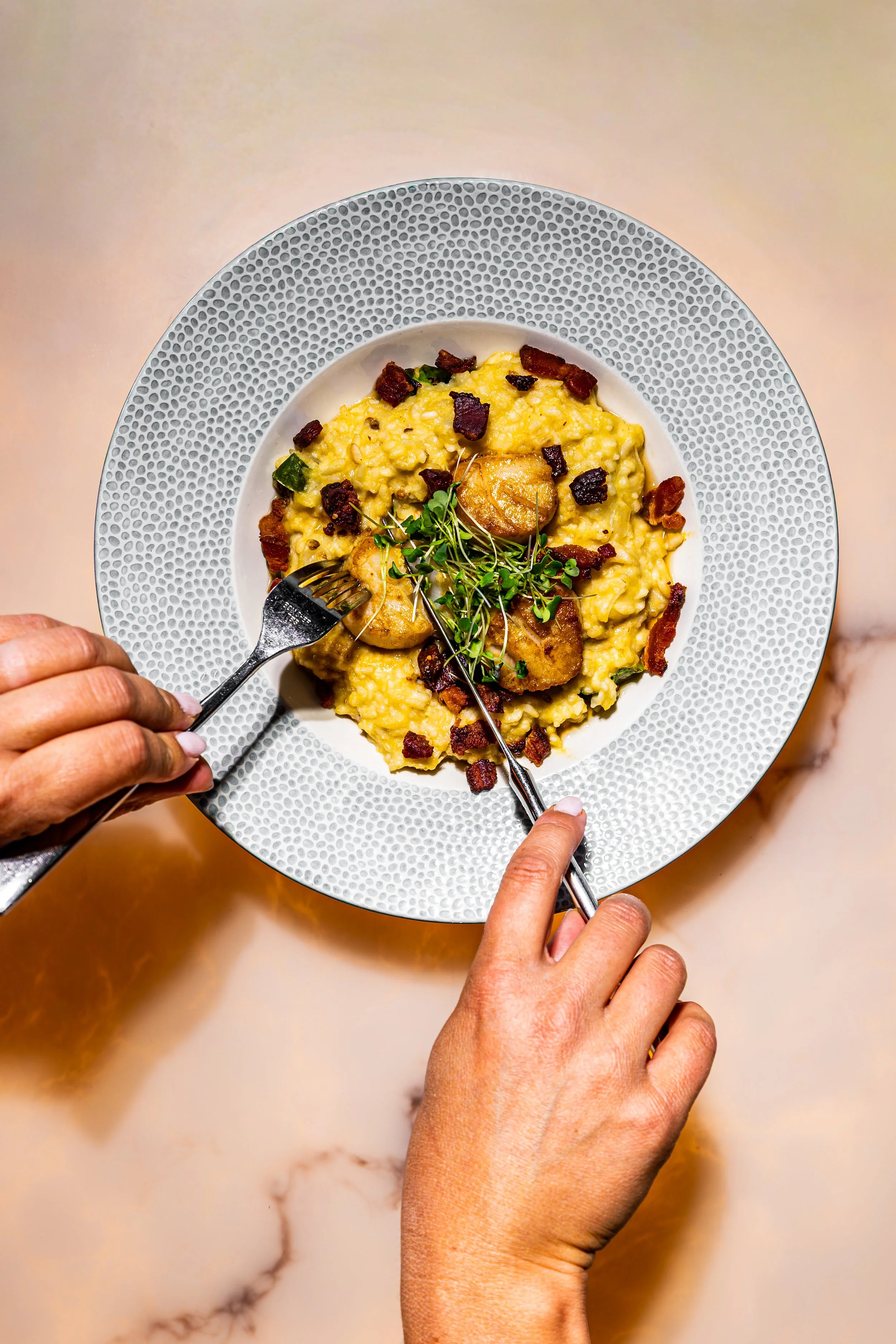 Top-down view of a person with tan skin holding a fork and knife, cutting into a plate of creamy risotto garnished with bacon bits, green herbs, and microgreens, on a white textured plate on a marble table.