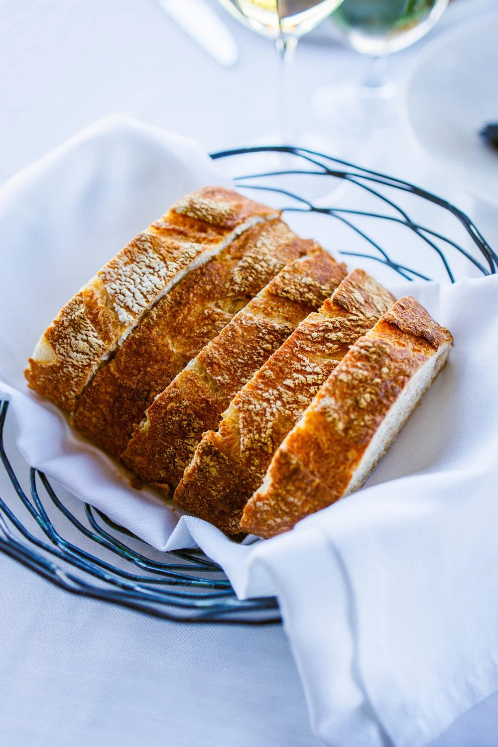 Sliced bread in a wire basket lined with white cloth, placed on a table with a white tablecloth, with a glass of wine in the background.