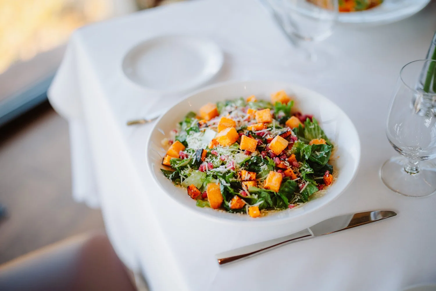 A bowl of mixed salad with chopped vegetables and shredded cheese on a white table, with a knife and empty condiment dish nearby.