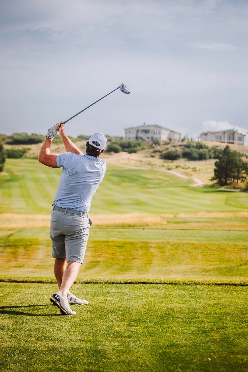 A man dressed in a light blue polo shirt, beige shorts, and a white cap is swinging a golf club on a golf course with grassy fairways and a few houses in the background.