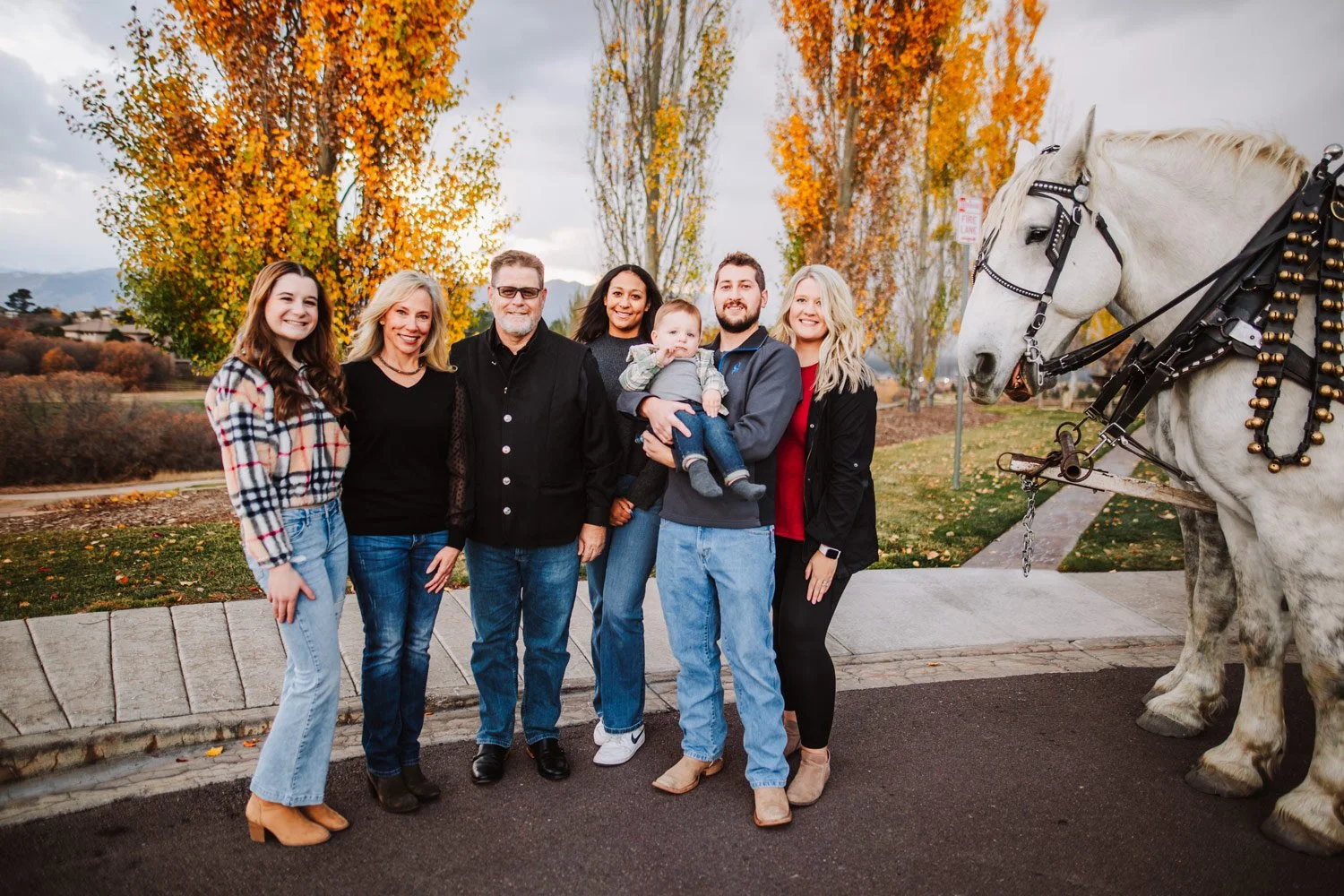 Family photo with six adults and one child standing outside during fall with trees and mountains in the background, along with a white horse on the right side.