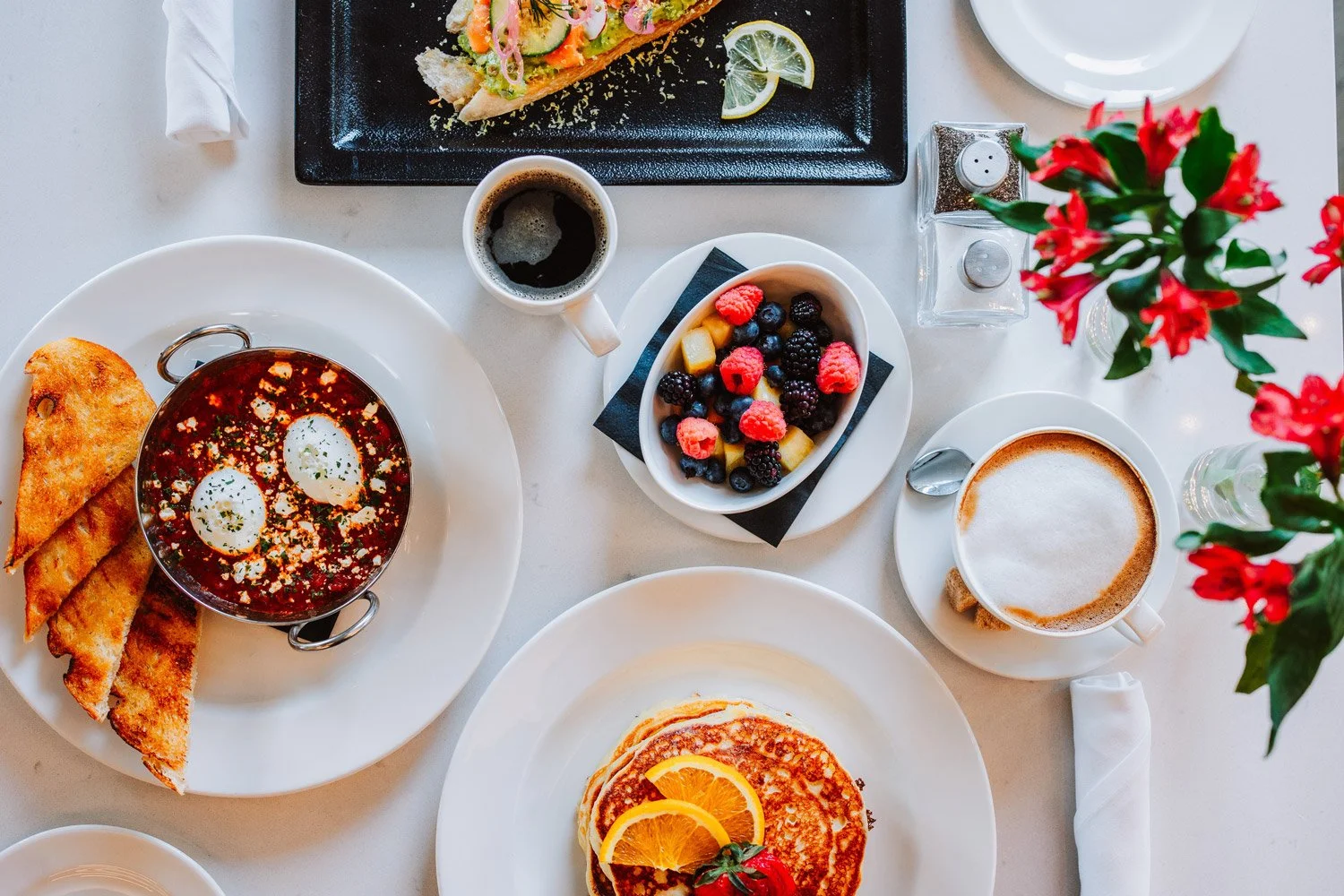 Breakfast spread with scrambled eggs topped with orange slices, a bowl of mixed berries, a cup of coffee with foam, a cup of black coffee, a side of toasted bread, and a plate with a vegetable dish garnished with lemon slices, set on a white table wi