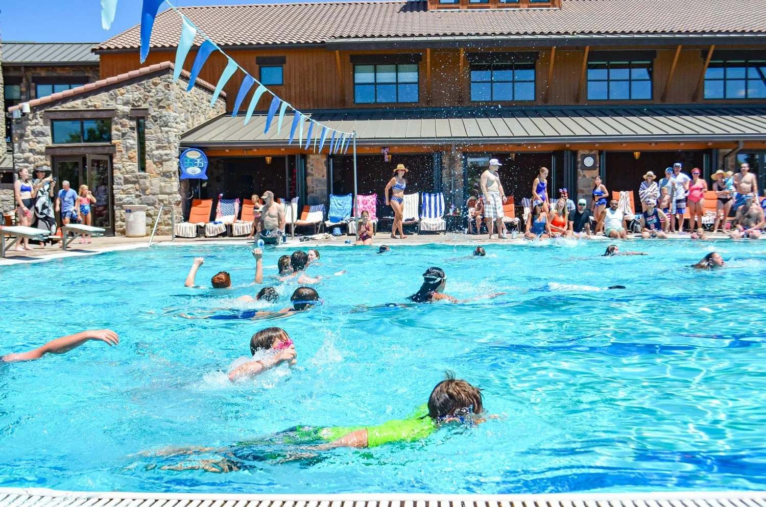A lively outdoor swimming pool scene with children and adults swimming and playing, surrounded by sunny weather and a stone and wood building with lounge chairs and people relaxing on the deck.