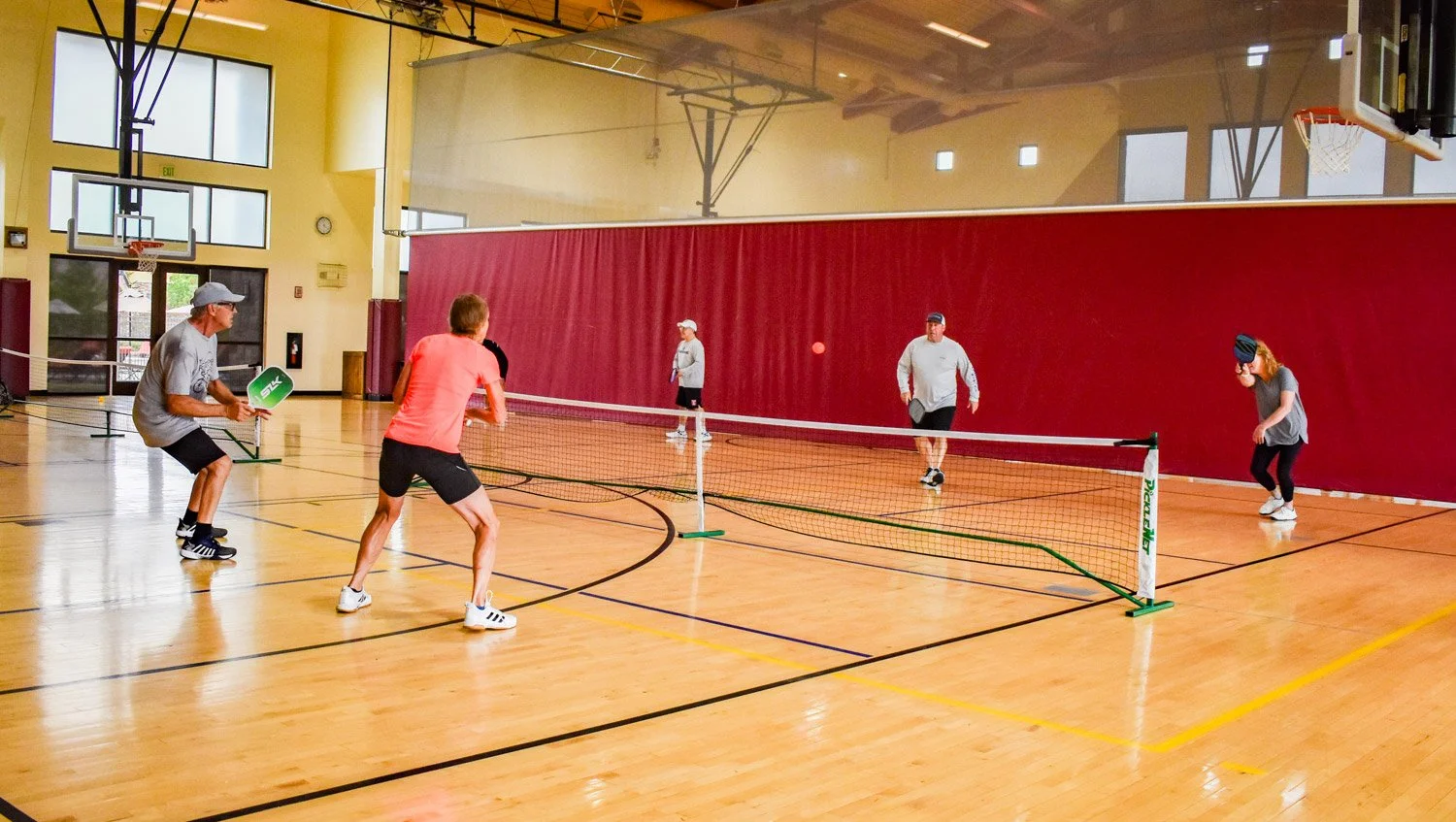 People playing pickleball indoors on a wooden court, with a net in the center, surrounded by yellow walls and windows, and a basketball hoop visible on the left side.