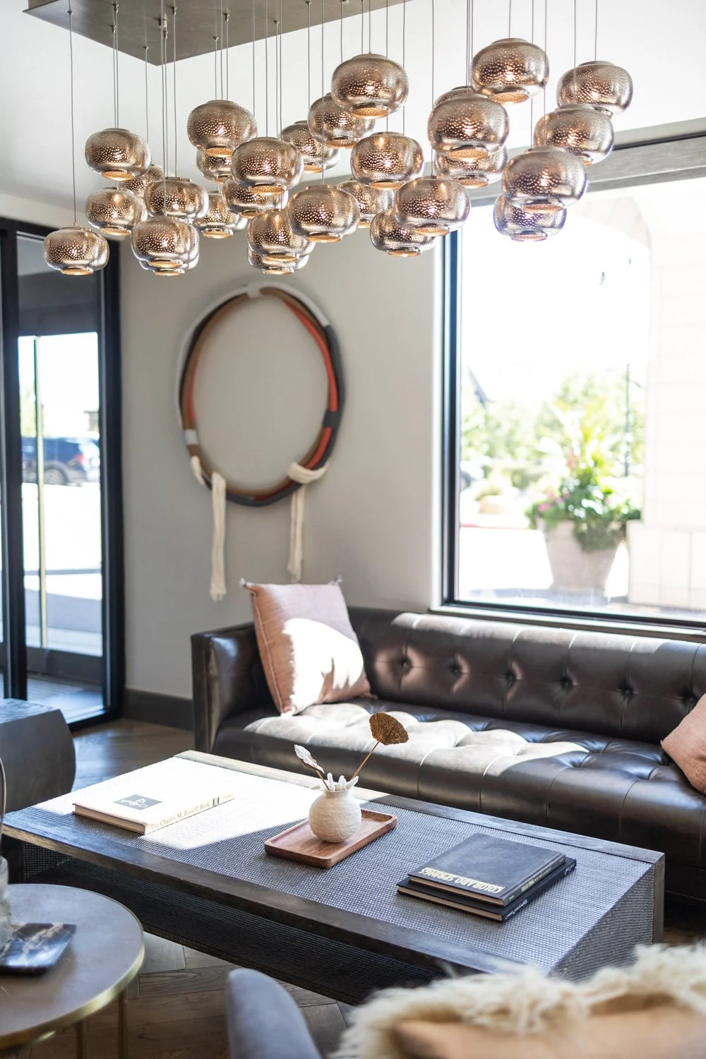 Contemporary living room with a black leather tufted sofa, a coffee table with books and a small vase with dried flowers, a large window with natural light, hanging metallic pendant lights, and wall decor featuring a circular woven piece.