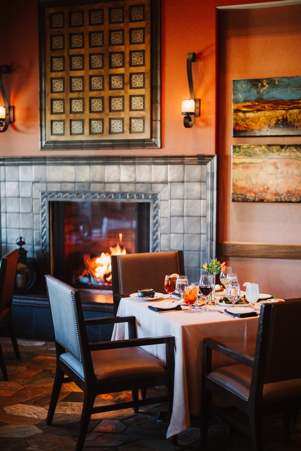 The Tack Room restaurant with a lit fireplace, a dining table set with wine glasses, water glasses, plates, and silverware, and decorated with a small flower arrangement. The room has warm orange walls, artwork, and wall-mounted lamps.