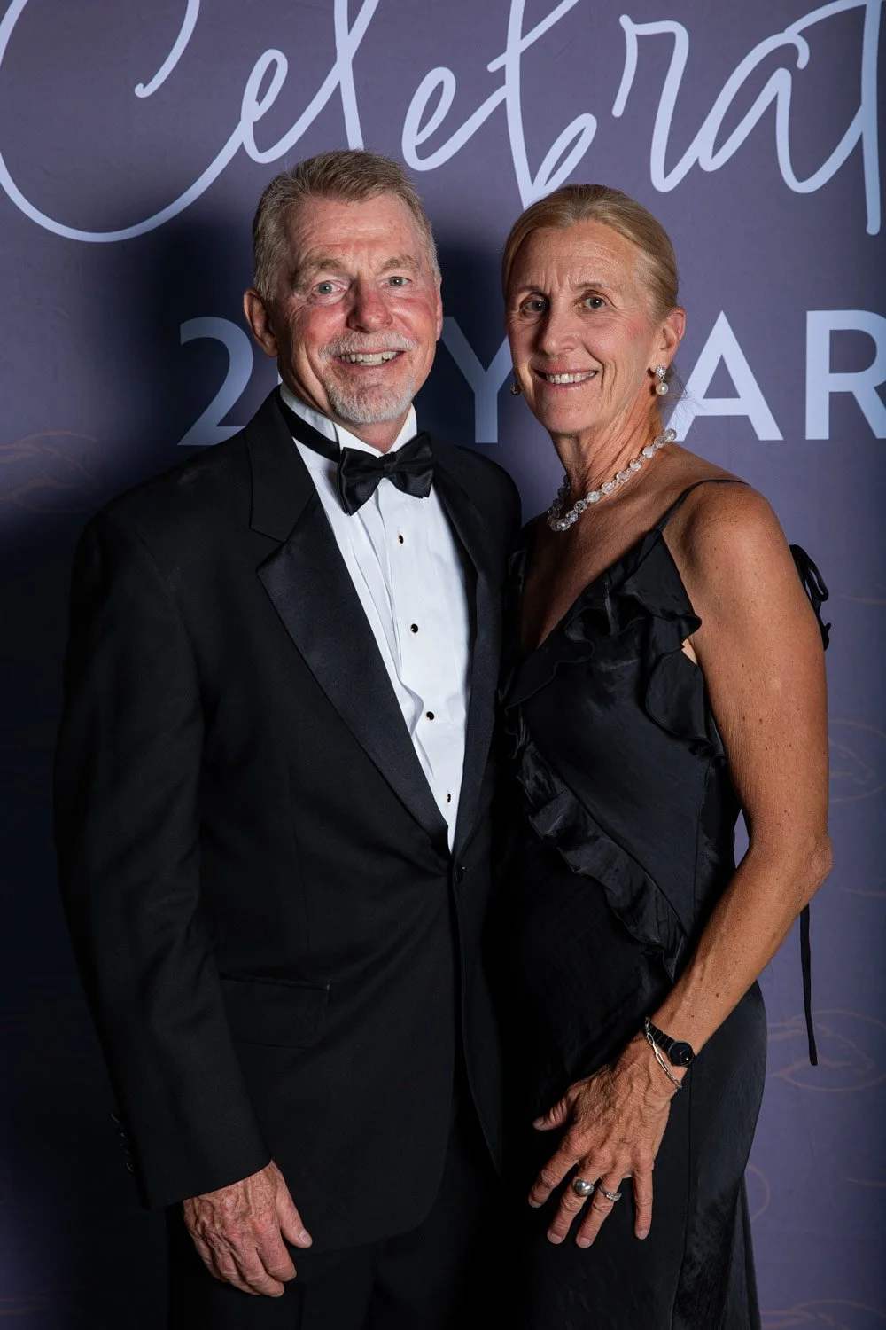 A smiling older man in a tuxedo and a smiling older woman in a black dress pose together at a formal event with a dark background that has elegant writing.