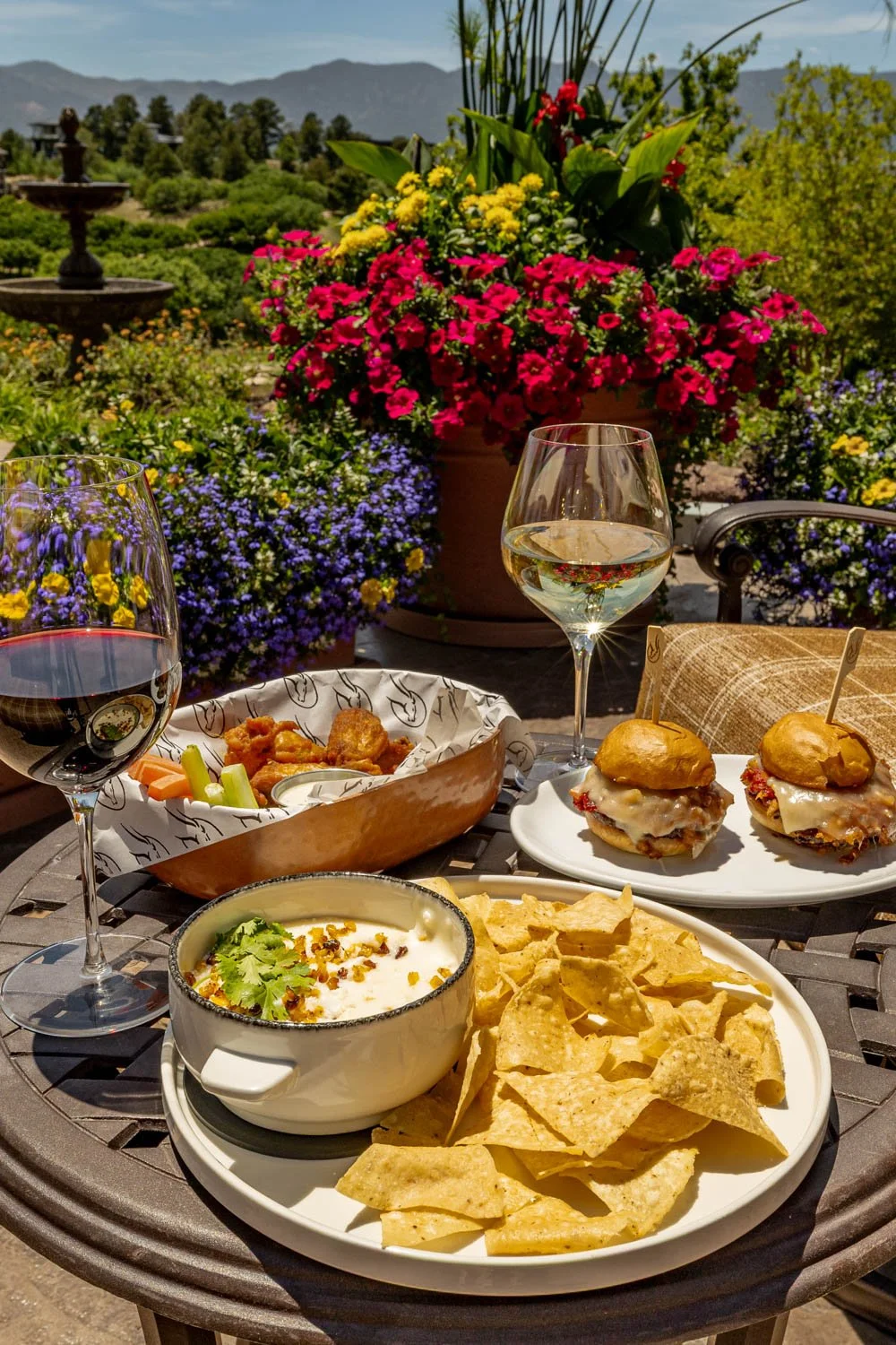 Outdoor dining table with food and drinks, including chips, salsa, sliders, chips and dip, and glasses of white wine, with colorful flowers and a mountain view in the background.