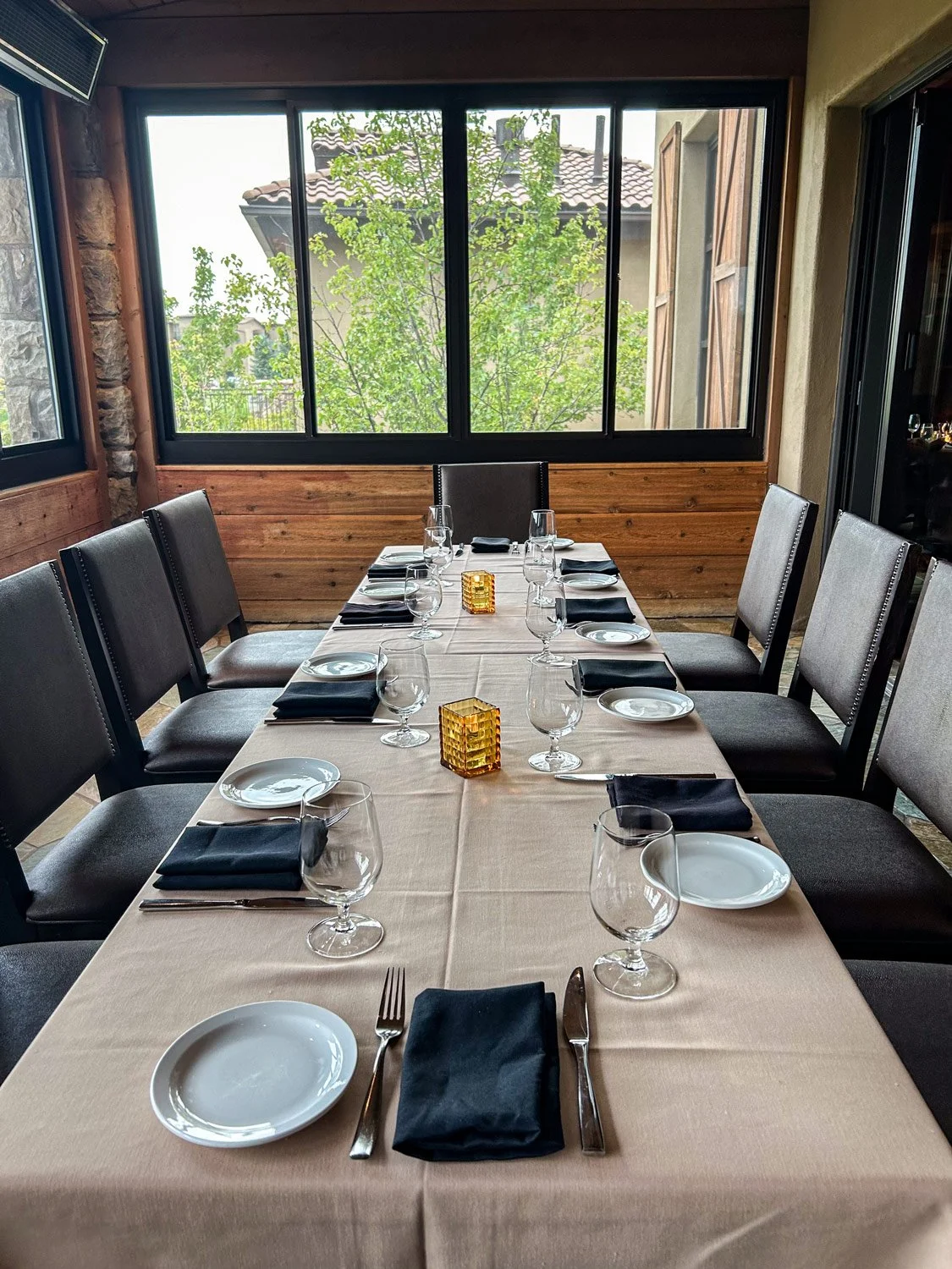 A dining table set for ten people with black napkins, plates, wine glasses, and two small amber candle holders, in a room with large windows and a view of trees and neighboring houses.