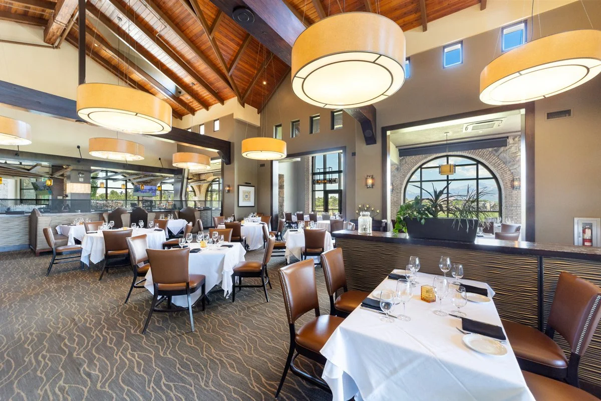 Elegant interior with white tablecloths, brown leather chairs, large circular pendant lights, and large windows showing a scenic view outside at The Steakhouse.