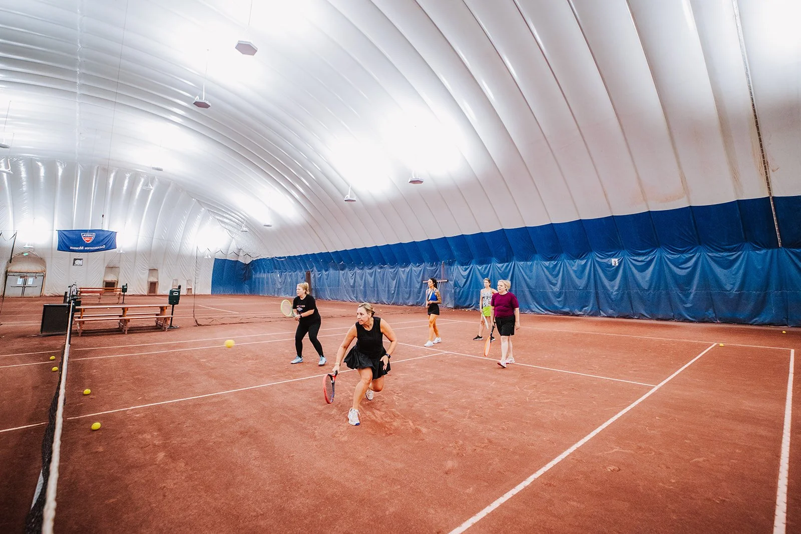 Women playing pickleball inside an indoor tennis court with a high, curved white ceiling and blue walls, some women are holding paddles and a yellow ball is visible on the court.
