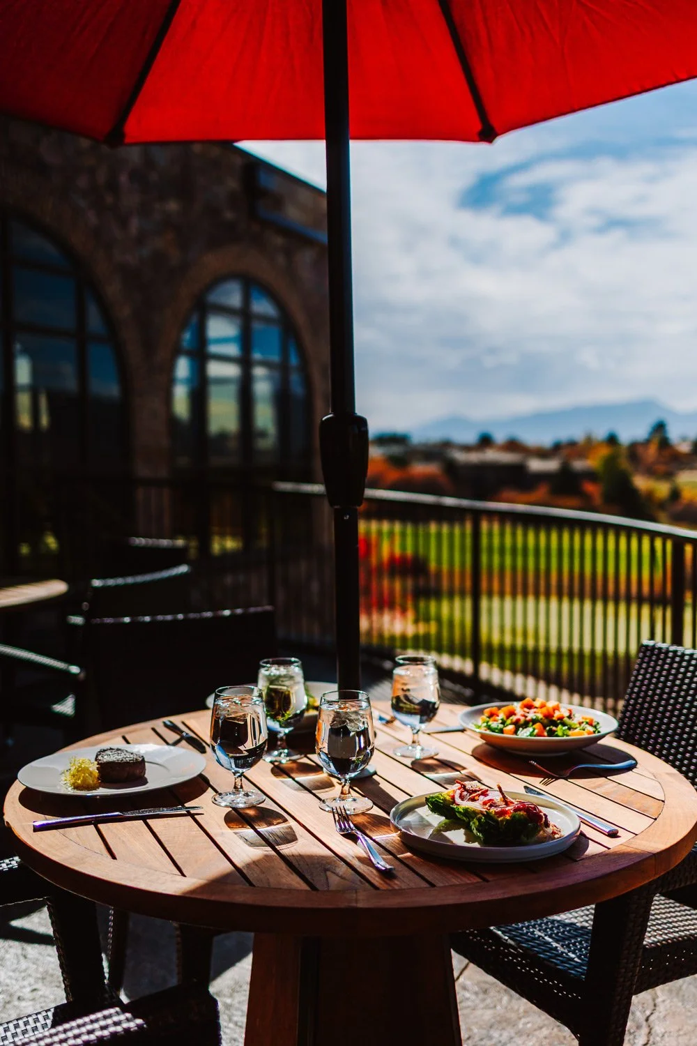 An outdoor dining table set with glasses of water, plates of food, and covered by a red umbrella on a balcony with a scenic mountain view.