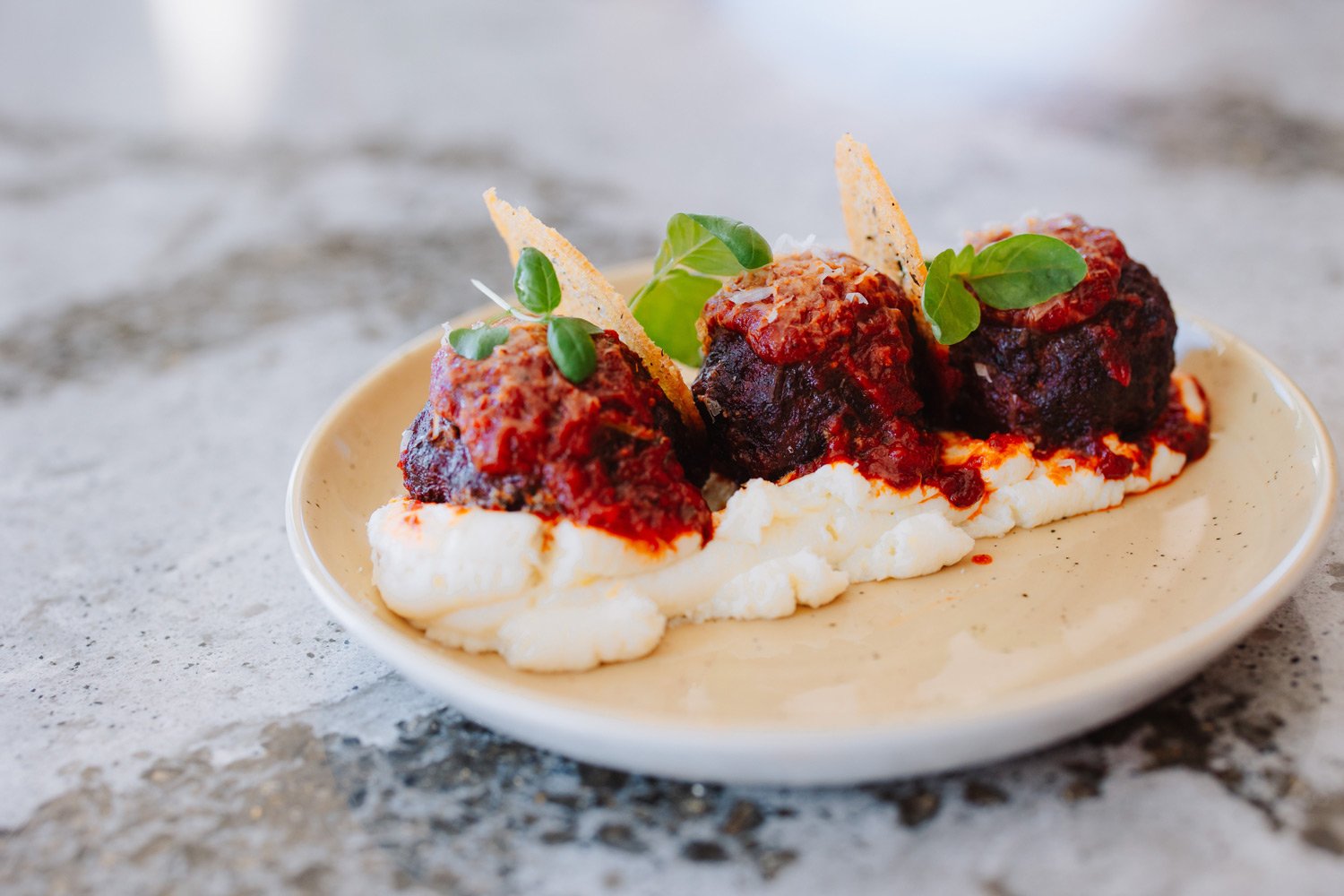 Plate of three meatballs topped with tomato sauce, garnished with basil leaves and Parmesan cheese, served over a bed of mashed potatoes or cream-based spread.